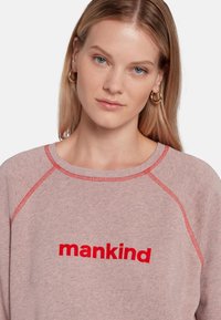 Sweatshirt rose clair avec broderie rouge "mankind", col côtelé et manches raglan, présentant une texture douce et des coutures contrastantes.