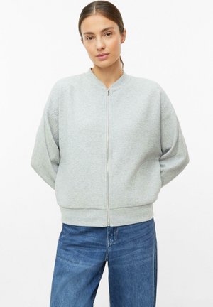 Frau mit braunen, zurückgebundenen Haaren, die einen hellgrauen Reißverschluss-Pullover und blaue Jeans vor einem schlichten weißen Hintergrund trägt.