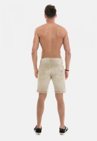 Shorts en denim beige avec ourlets retroussés, taille mi-haute et poches arrière. Portés avec des baskets noires, modèle tourné le dos à l'appareil photo.