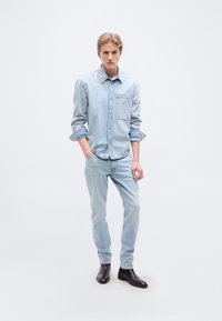 Φωτεινό γαλάζιο πουκάμισο από τζιν με τσέπη στο στήθος, συνδυασμένο με ανοιχτόχρωμα γαλάζια τζιν. Η εμφάνιση ολοκληρώνεται με σκούρα καφέ μποτάκια έως τον αστράγαλο.