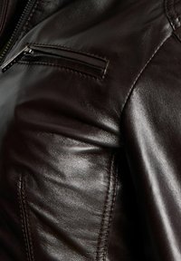 Zoom na temno rjavo usnjeno jakno, ki prikazuje žep na zadrgo in podrobno obšito na rami in prsnem področju.