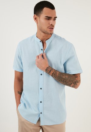 Camicia azzurra a maniche corte con colletto alla cinese e bottoni neri, caratterizzata da un tessuto texturizzato e polsini arrotolati.