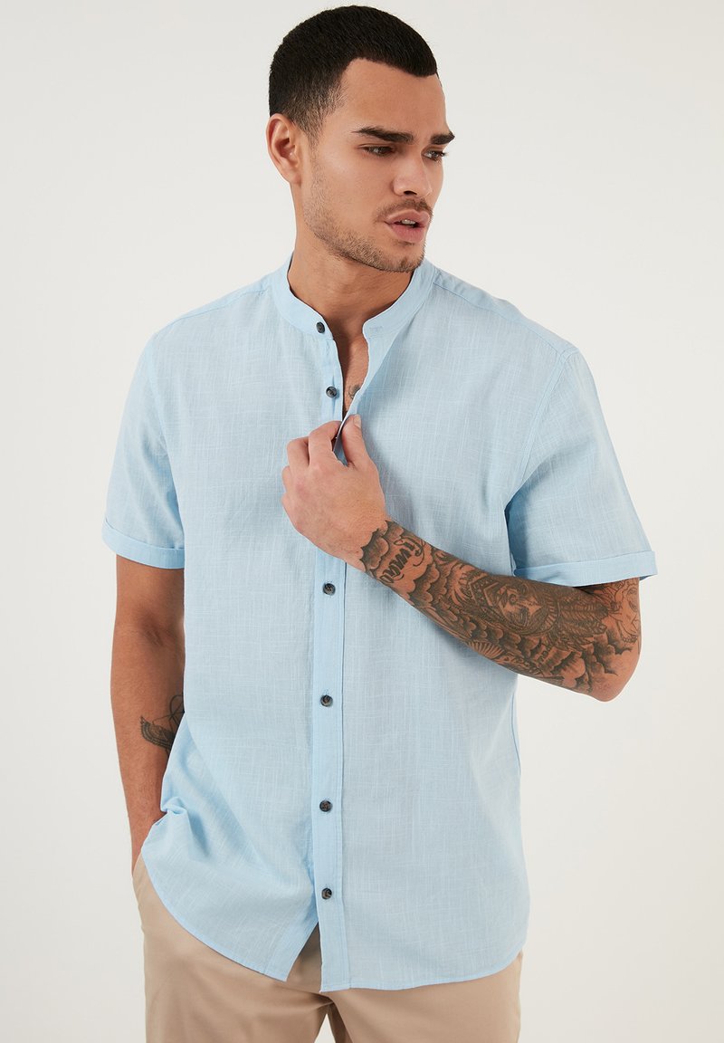 Camisa de manga corta de color azul claro con cuello mao y botones negros, con una tela texturizada y puños enrollados.