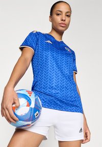 Blue patterned soccer jersey with gold accents, paired with white shorts. The player holds a multicolored soccer ball in one hand.