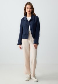 Navy linnen cropped jack met een knoopsluiting, twee voorzakken, gedragen over een witte top en gecombineerd met lichtbeige wijde broek en sneakers.