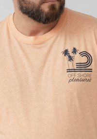 Hellblaues T-Shirt mit einem gedruckten Design von Palmen und dem Text "OFF SHORE pleasures" in Dunkelblau, mit einem runden Ausschnitt.