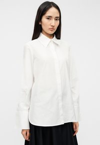 Άσπρο πουκάμισο με μακριά μανίκια, μυτερό γιακά, κουμπιά μπροστά και φαρδιά μανσέτα, κατασκευασμένο από λεία, ελαφριά ύφασμα.