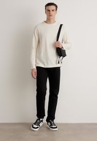 Lys kremfarget strikkegenser, svarte bukser, holder en liten svart veske. Svart og hvit sneakers, står mot en nøytral bakgrunn.