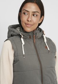 Grahamannte Weste mit Kapuze, ausgestattet mit einem Frontreißverschluss, zwei weißen Kordeln und braunen Akzenten. Über einem hellen Pullover getragen.