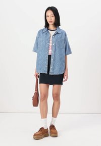 Cămașă oversized din denim de culoare albăstruie, cu două buzunare în față, purtată peste un tricou cu grafică. Asortată cu o fustă mini neagră și adidași cu talpă înaltă.