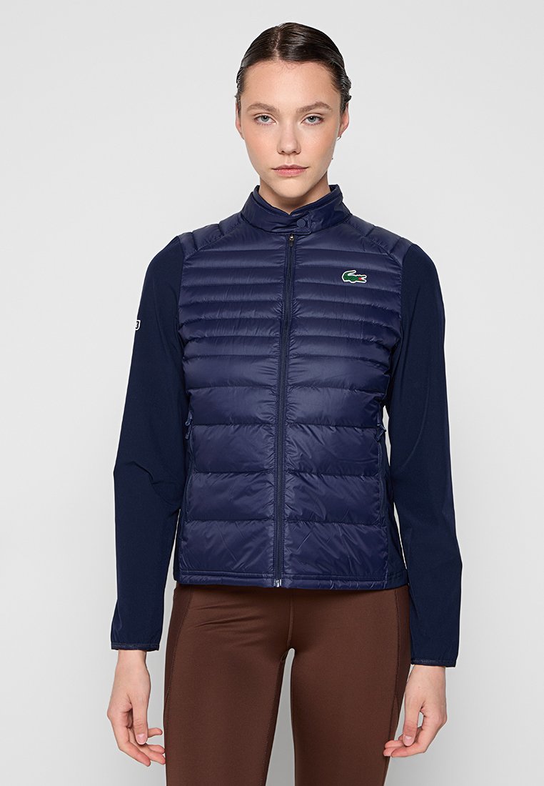 Doudoune sans manches bleu marine avec un col haut, présentant des motifs matelassés horizontaux, une fermeture éclair, et un petit logo vert Lacoste sur la poitrine.