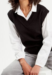 Pull en tricot noir sans manches avec encolure en V, porté sur une chemise blanche à manches longues. Un pantalon gris est partiellement visible. Couleurs unies, tissu texturé.