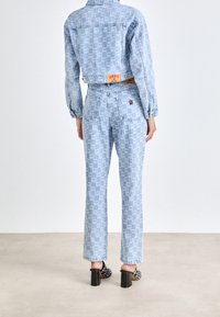 Ljusblå denimjacka och byxset med upprepande logomönster, kontrasterande orange etiketter och tydliga svarta högklackade skor.