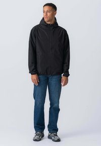 Sort letvægtsjakke med hætte, lynlås lukning og elastiske manchetter; sat sammen med blå jeans og grå træningssko. Simpelt design, minimale detaljer.