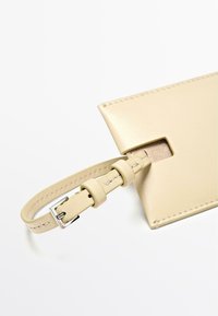 Beige leren kaarthouder met een gladde textuur, voorzien van een uitsparing voor gemakkelijke toegang en een verstelbare band met een zilveren gesp.