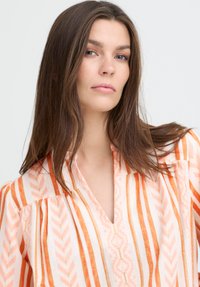 Blouse ample à rayures verticales orange et rose. Présente un col en V et un tissu texturé avec un motif géométrique subtil.