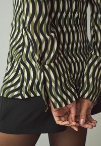 Blusa verde e crema con motivo a onda, maniche lunghe, taglio aderente e dettagli con bottoni ai polsini, abbinata a una gonna nera.