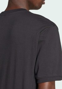 Černé bavlněné tričko s kulatým výstřihem, krátkými rukávy a viditelným švem. Hladká textura a volný střih, zdůrazňující detail ramen.