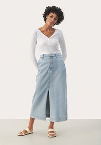 Jupon maxi en denim bleu clair avec une fente avant, taille haute et poches au dos. Associé à un top ajusté à manches longues blanc et des sandales.