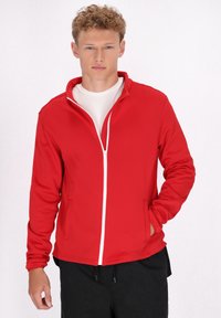Rode zip-up jas met een staande kraag, gemaakt van gladde stof, voorzien van een witte rits en twee zijzakken. Geshould met een wit shirt eronder.