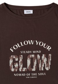 T-shirt marron avec le texte "GLOW" en sequins, et les phrases imprimées "FOLLOW YOUR", "STEADY MIND", "NOMAD OF THE SOUL" et "STAY CURIOUS".