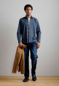 Καρό πουκάμισο με κουμπιά σε σκούρο μπλε, ανοιχτόχρωμο μπλουζάκι από κάτω, τζιν παντελόνι, κρατώντας μια καφέ ζακέτα. Φορεμένο με μαύρα παπούτσια.