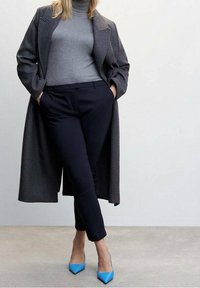 Manteau en laine gris porté sur un col roulé gris, associé à un pantalon bleu marine et des escarpins bleu vif pointus, créant un contraste de couleurs équilibré.