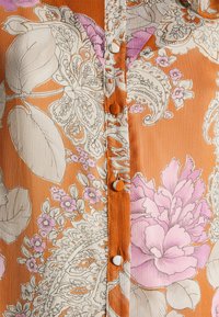 Tessuto arancione con dettagliati motivi floreali e paisley bianchi e rosa, con bottoni rivestiti lungo una cucitura centrale.