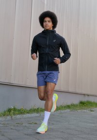 Chaqueta de running ligera negra, pantalones cortos azules, calcetines blancos y zapatillas de running amarillas neón. El modelo corre por una superficie pavimentada.