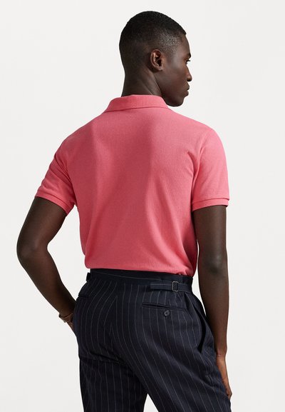 Camiseta polo vermelha com mangas curtas, fabricada em tecido texturizado, vista traseira, usada com calças azul-marinho com riscas verticais, apresentando bolsos laterais e passantes para cinto.