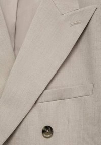 Giacca su misura beige chiaro con rever affilati, tessuto testurizzato e chiusura a un bottone. Include un taschino laterale.