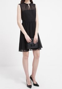 Czarna sukienka bez rękawów z przezroczystą górną częścią, zbieranym wcięciem w talii i zwiewnym dołem. Połączona z czarną torebką i spiczastymi szpilkami.