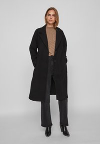 Γυναίκα που στέκεται με τα χέρια στις τσέπες, φορώντας έν�α μαύρο μακρύ παλτό, καφέ με καρό μπλουζάκι, μαύρα τζιν και μαύρες μπότες με τακούνι σε γκρίζο φόντο.