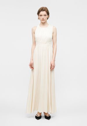 Donna con capelli corti che indossa un abito crema senza maniche, lungo fino a terra, e scarpe nere con punta appuntita, in piedi davanti a uno sfondo bianco semplice.