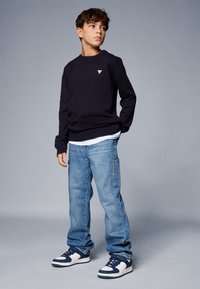 Sweatshirt bleu marine avec un petit logo triangulaire, associé à un jean baggy bleu clair avec des poches latérales et des baskets bleu marine avec des accents blancs.