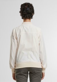 Lys beige bomberjakke med ribbestrikkede mansjetter og krage, med en glatt, noe glinsende tekstur og enkelt design. Synlige sømdetaljer.