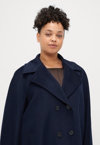 Cappotto navy doppiopetto con ampi rever, bottoni neri e tasche laterali. Texture liscia, vestibilità sartoriale e dettagli minimi.