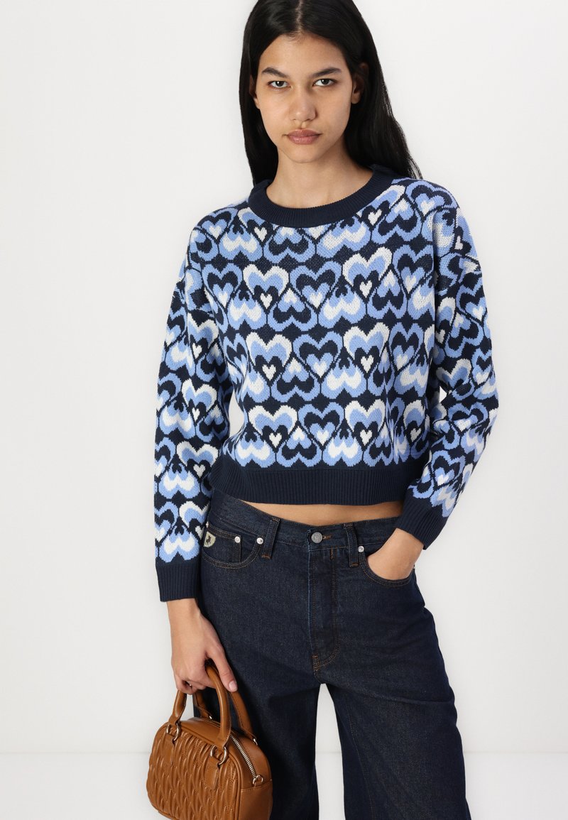 Gebreide trui in blauwe tinten met een hartpatroon, ronde halslijn en geribbelde zoom. Gecombineerd met donkere spijkerbroek en een bruine handtas.