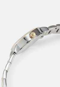 Ceas de mână cu tonuri de argint și aur, având un cadran rotund, brățară din metal polisat și o coroana proeminentă pe lateral.