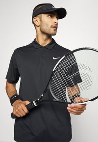 Schwarzes Nike-Polohemd mit Kragen und Logo, kombiniert mit einem schwarzen Handgelenkband und hält einen Tennisschläger mit strukturiertem Griff.