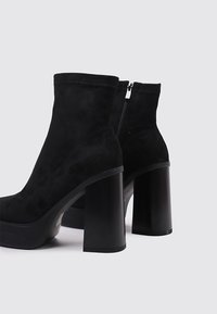 Botas de tobillo de ante negro con un tacón bloque negro brillante, que cuentan con un cierre de cremallera en la parte posterior y una superficie superior suave y texturizada.