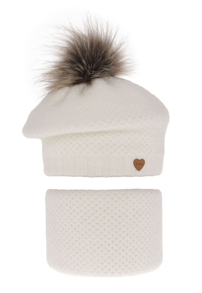 Cappellino bianco lavorato a maglia con pompon in pelliccia sintetica e piccolo etichetta a forma di cuore marrone. Sciarpa coordinata bianca in maglia sotto, superficie testurizzata.