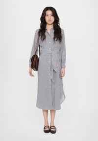 Abito a righe blu navy e bianco, di lunghezza media con maniche lunghe, vita cintrata e spacco laterale. Abbinato a una borsa a tracolla marrone e scarpe.