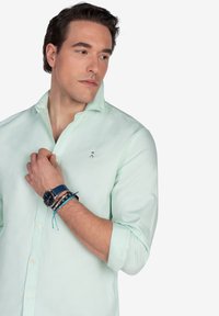 Camisa verde claro de manga larga, con cuello abotonado y un pequeño logotipo bordado. El modelo lleva varias pulseras en la muñeca y un reloj oscuro.