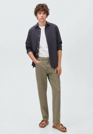 Camicia button-up blu scuro, t-shirt bianca, pantaloni verde oliva e sandali marroni. Il modello sta dritto su uno sfondo chiaro.