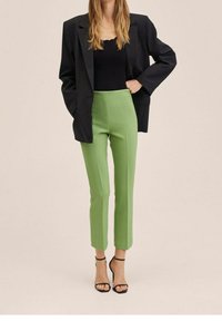 Vrouw in een zwarte blazer, zwart top, lichtgroene nauwsluitende broek en zwarte enkelbandhakken, staande met één hand op de heup.