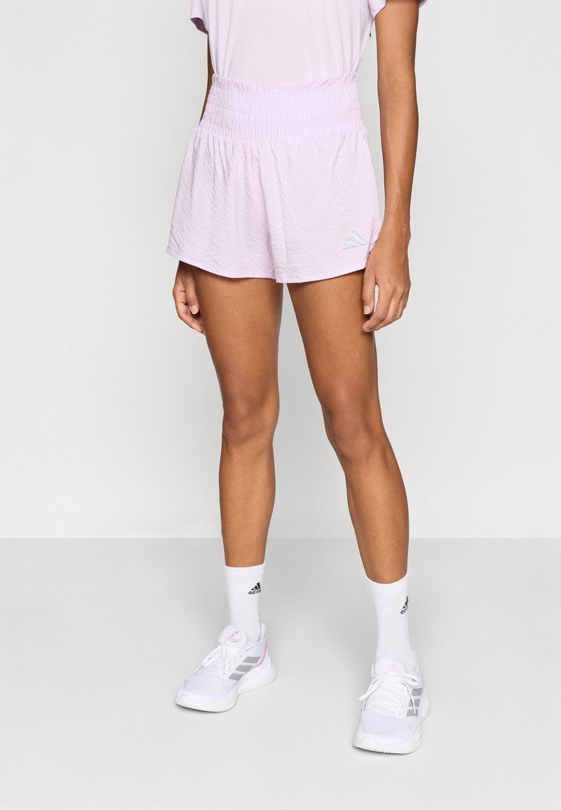 Gaiši rozā šorti ar elastīgu jostu, vieglu audumu un nelielu zilu logotipu. Kopā valkāti ar baltām sporta kurpēm un zeķēm.