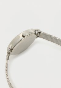 Relógio com bracelete em malha prateada e caixa redonda polida em prata. A vista lateral mostra uma coroa para ajustes e um acabamento metálico suave.