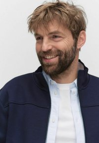 Marinblå zip-up jacka över en ljusblå randig skjorta. Jackan har en hög krage och en slät textur, medan skjortan har knapprad.