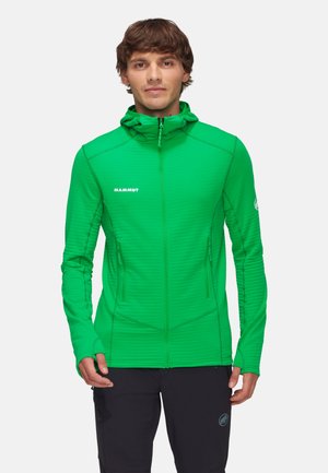 Ung mand iført en lys grøn lynlåshættejakke med "Mammut"-logo, sorte bukser, stående med hænderne afslappet ved siderne.